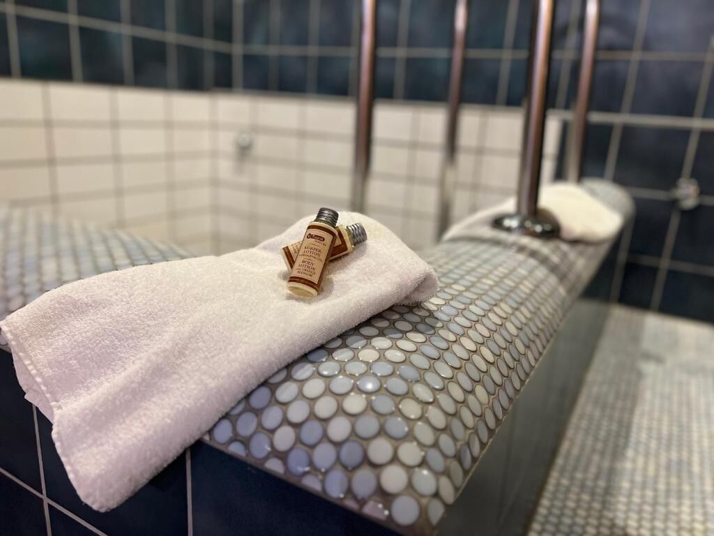 Un baño con piso de cerámica, una toalla blanca y una botella de loción sobre una banca.