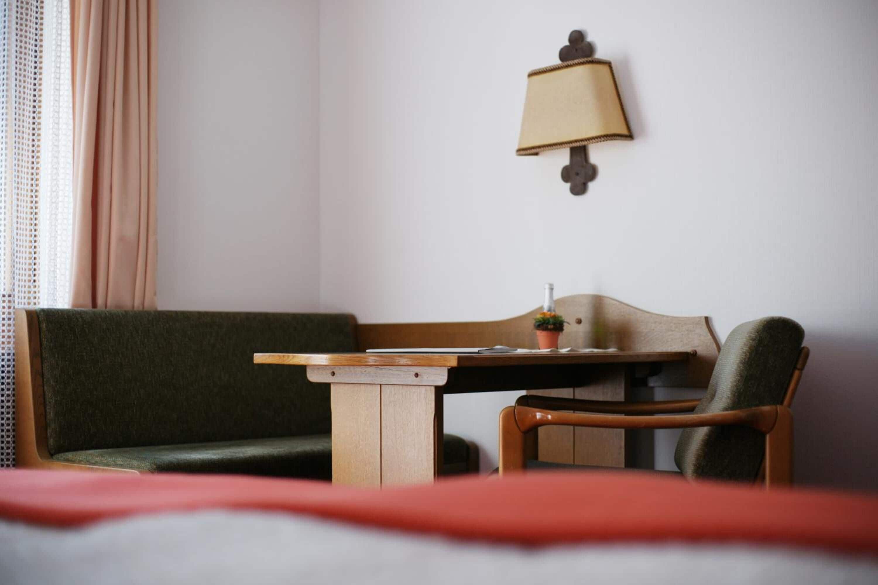 A hotel room with a bed, a wooden table, a green sofa, a wooden chair, and a wall-mounted lamp.