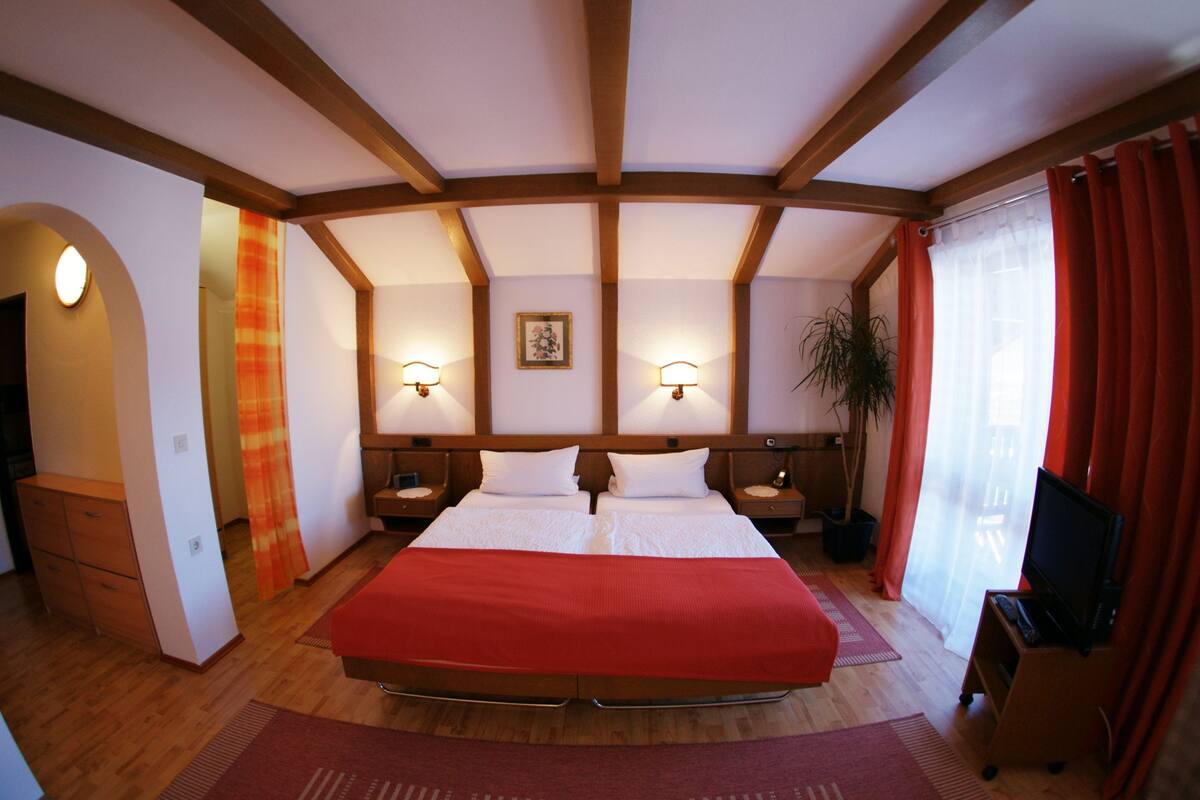 Une chambre d’hôtel avec un grand lit, des poutres apparentes en bois au plafond, deux tables de chevet avec des lampes, un meuble pour la télévision et une plante en pot.