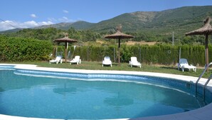 Ein Schwimmbecken mit Liegestühlen und Reetdach-Sonnenschirmen, umgeben von Grünflächen und Bergen.