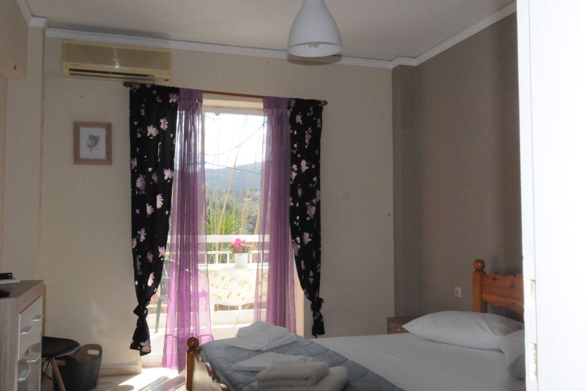 Un dormitorio con cama, ventana con cortinas moradas, cabecera de madera y lámpara blanca.