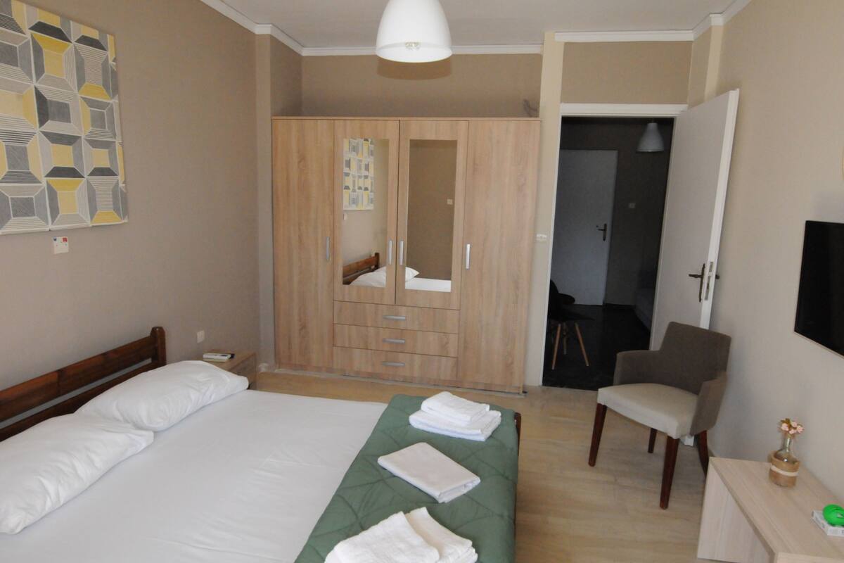 Un dormitorio con cama, colcha verde, armario de madera, una silla y televisor en la pared.