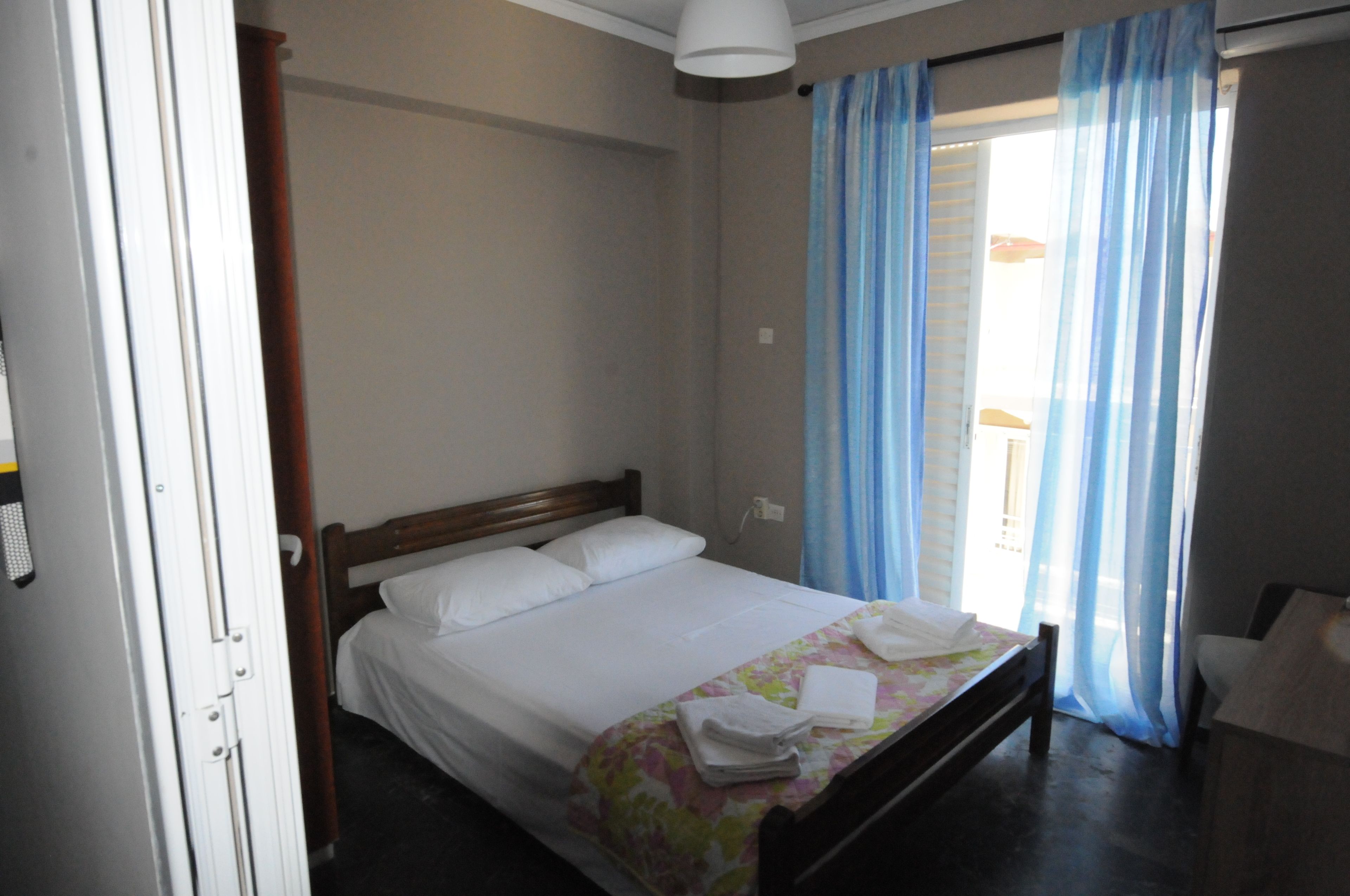 A single bed with white linens, a wooden headboard, and a floral-patterned bedspread. There is a window with blue curtains, and a white ceiling light is visible.