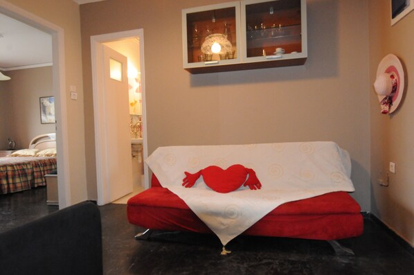 A cozy room with a bed, a red sofa, a small table, and a wall-mounted shelf with decorative items. - Kinetta Apartments (Oropos)