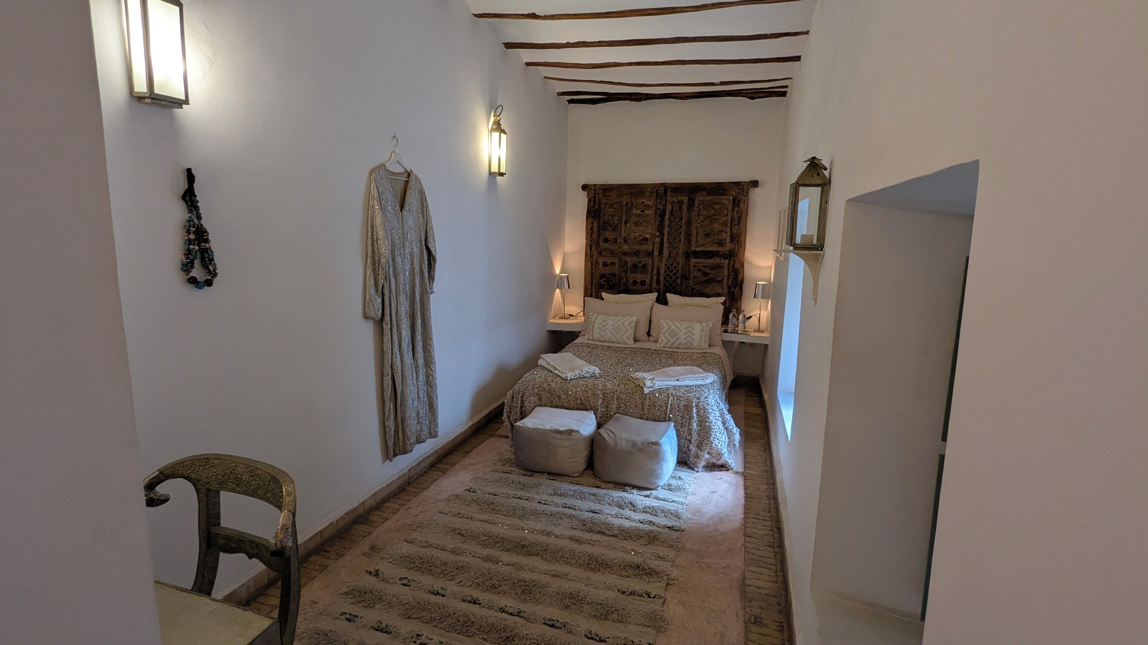 A narrow hallway leading to a bedroom with a bed, two bedside tables, a lamp, and a hanging garment.