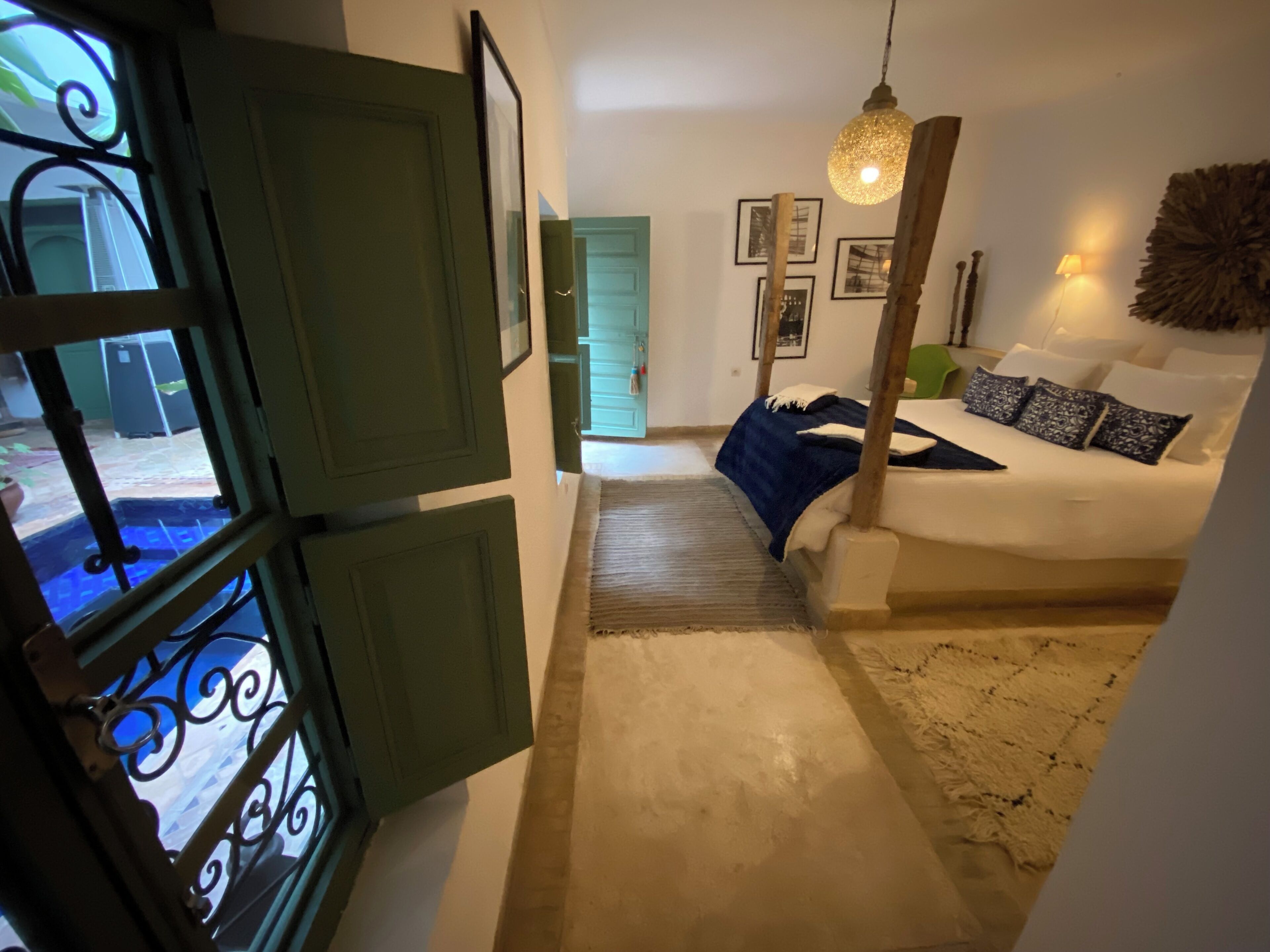 Un dormitorio con una cama, cabecera de madera, lámpara colgante y vistas al exterior a través de una ventana con contraventanas verdes.