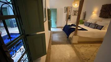 Un dormitorio con una cama, cabecera de madera, lámpara colgante y vistas al exterior a través de una ventana con contraventanas verdes.
