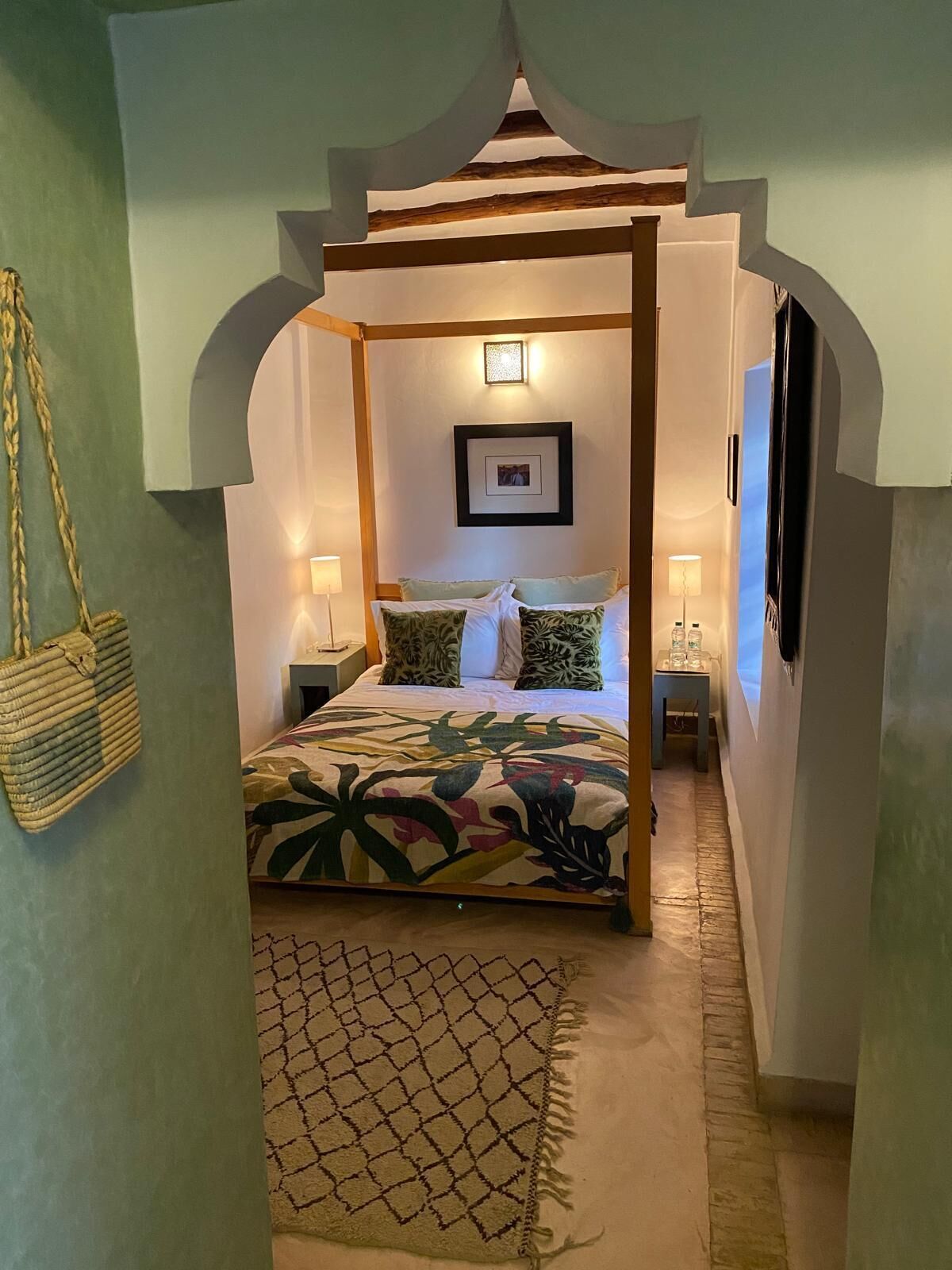 A bedroom with a canopy bed, a framed picture on the wall, and a woven bag hanging on the wall.