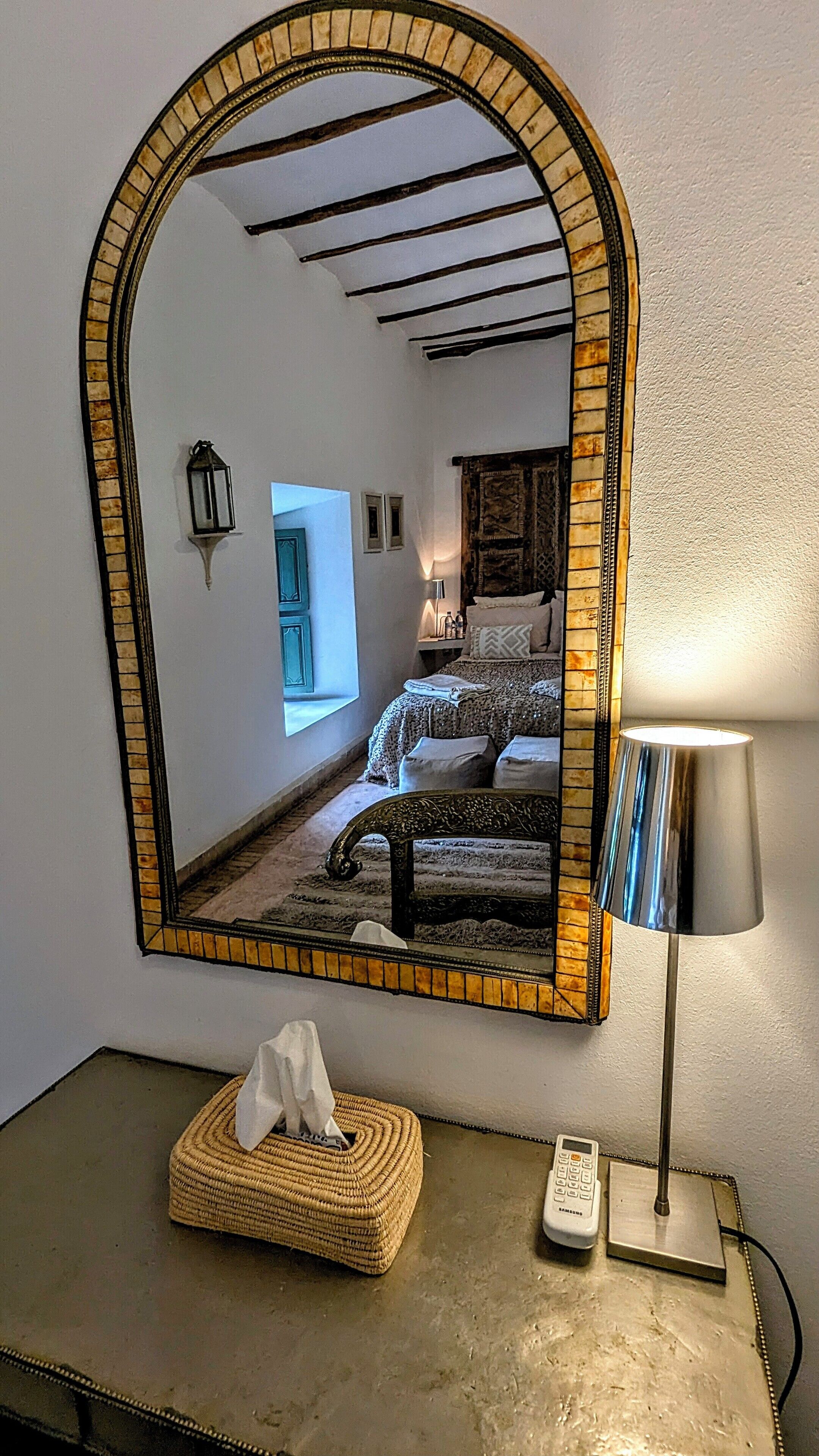 A mirror with a mosaic frame reflecting a bedroom with a bed, a bedside table, and a lamp.