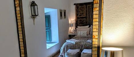 A mirror with a mosaic frame reflecting a bedroom with a bed, a bedside table, and a lamp.