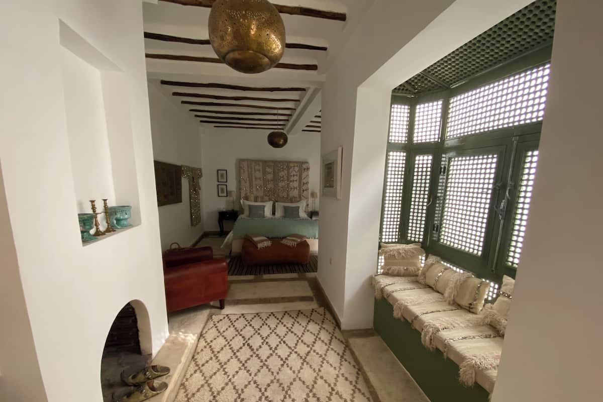 Una habitación con alfombra estampada, una cama con cabecera decorativa, un banco con cojines, una lámpara colgante y una ventana con diseño de cuadrícula.