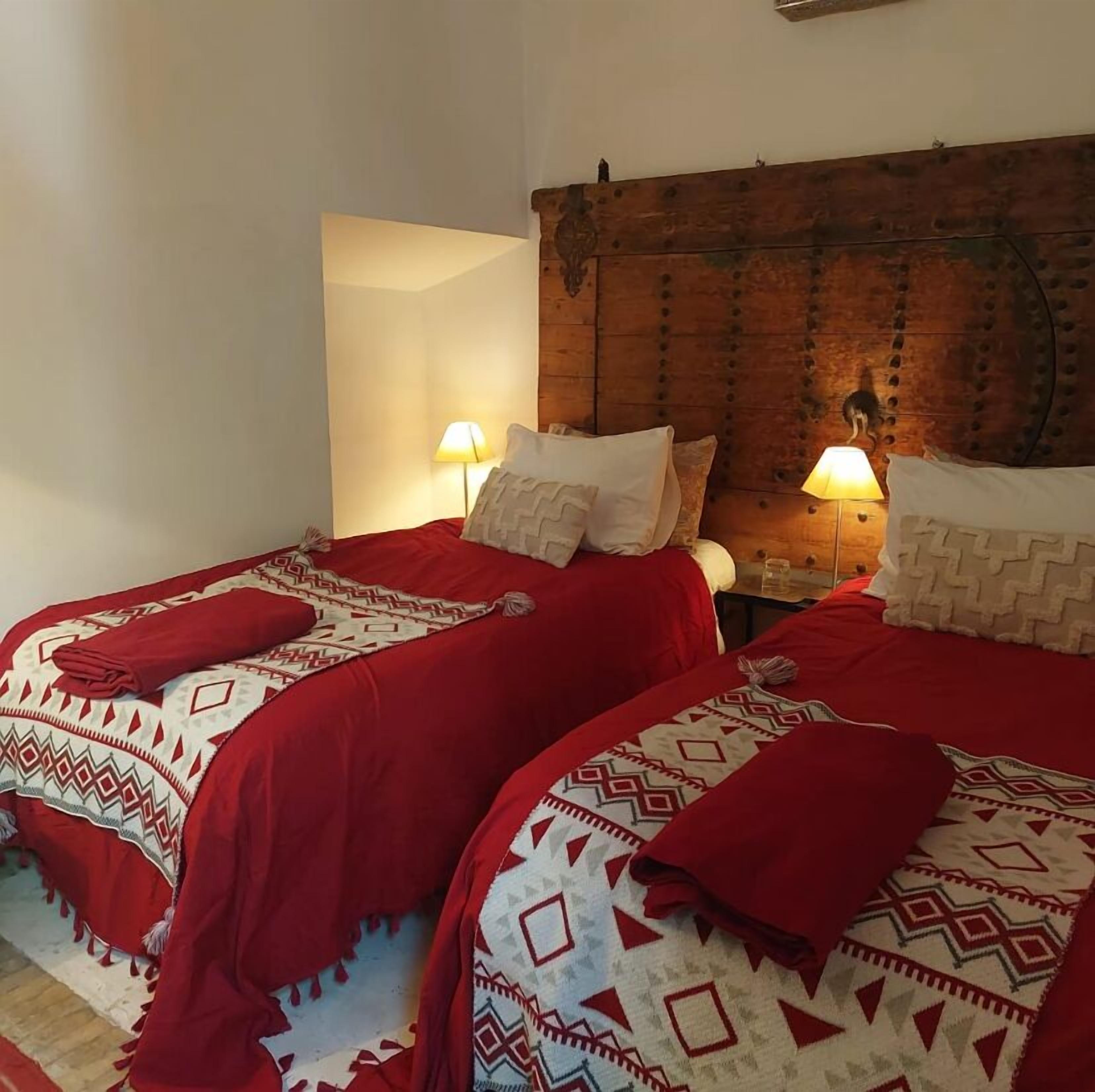 Quarto com duas camas, cada uma com roupa de cama e almofadas estampadas, cabeceira de madeira e duas luminárias de mesa.