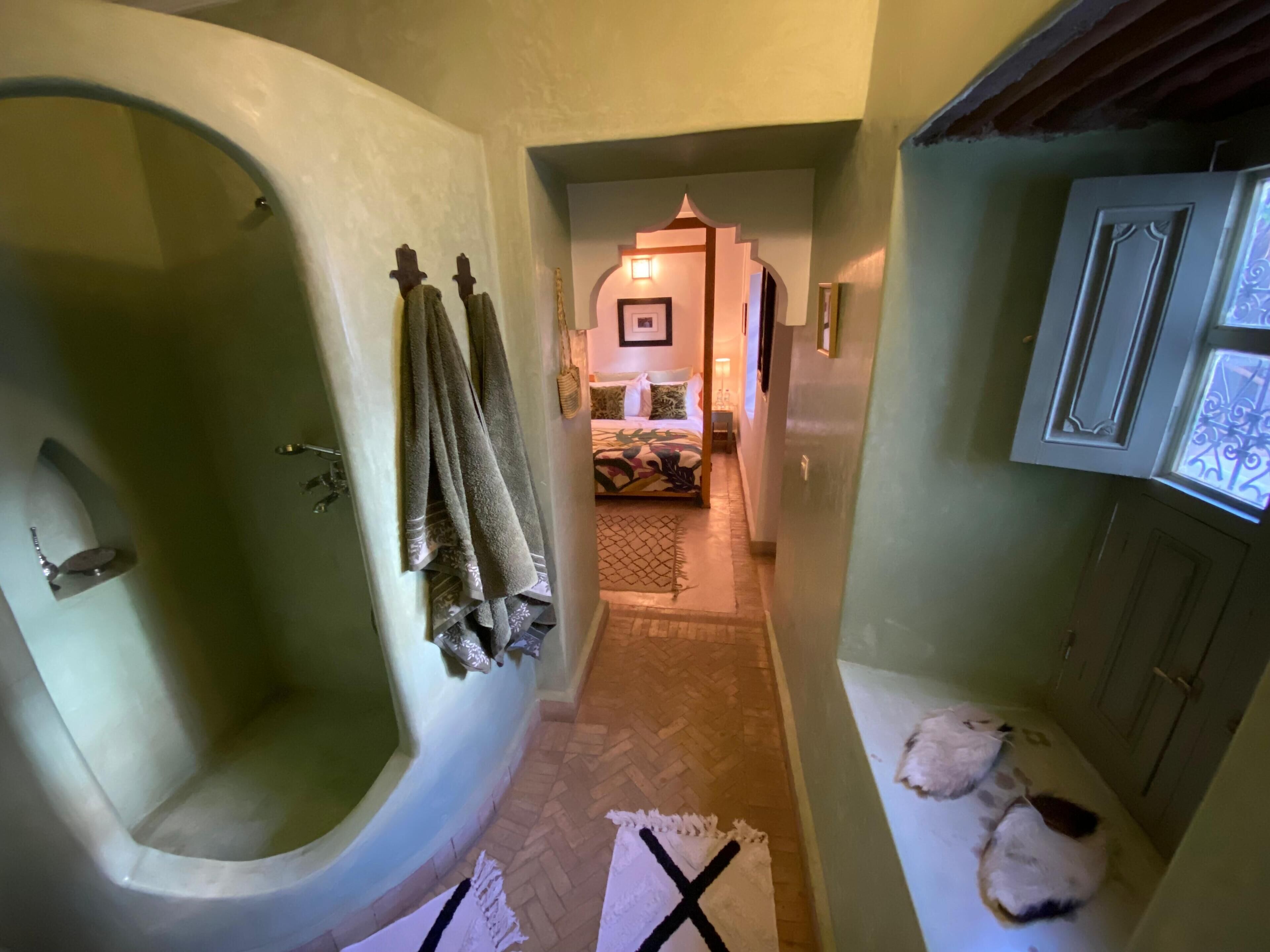 A narrow hallway with a large bathtub on the left, a bed with patterned bedding in the center, and a cabinet with a window on the right.
