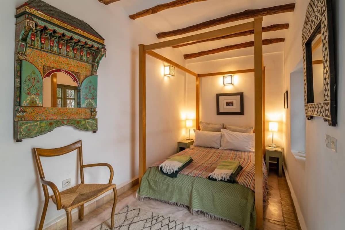 Un dormitorio con cama con dosel, una silla de madera, un espejo decorativo y un tapete estampado.