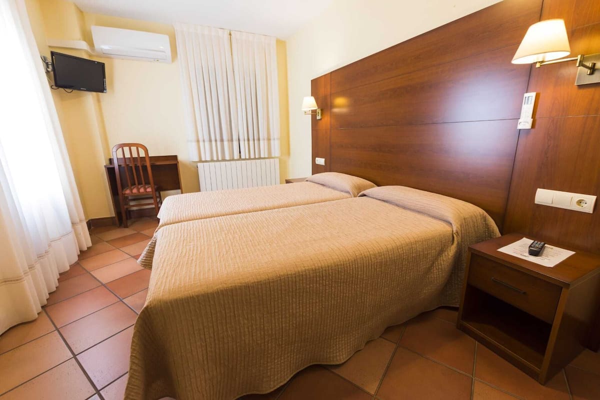 A hotel room with two beds, a wooden headboard, a bedside table with a lamp, a chair, and a TV mounted on the wall.