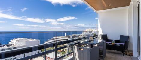 Un balcón con vistas al mar y edificios, amueblado con sillas de mimbre y una mesa.