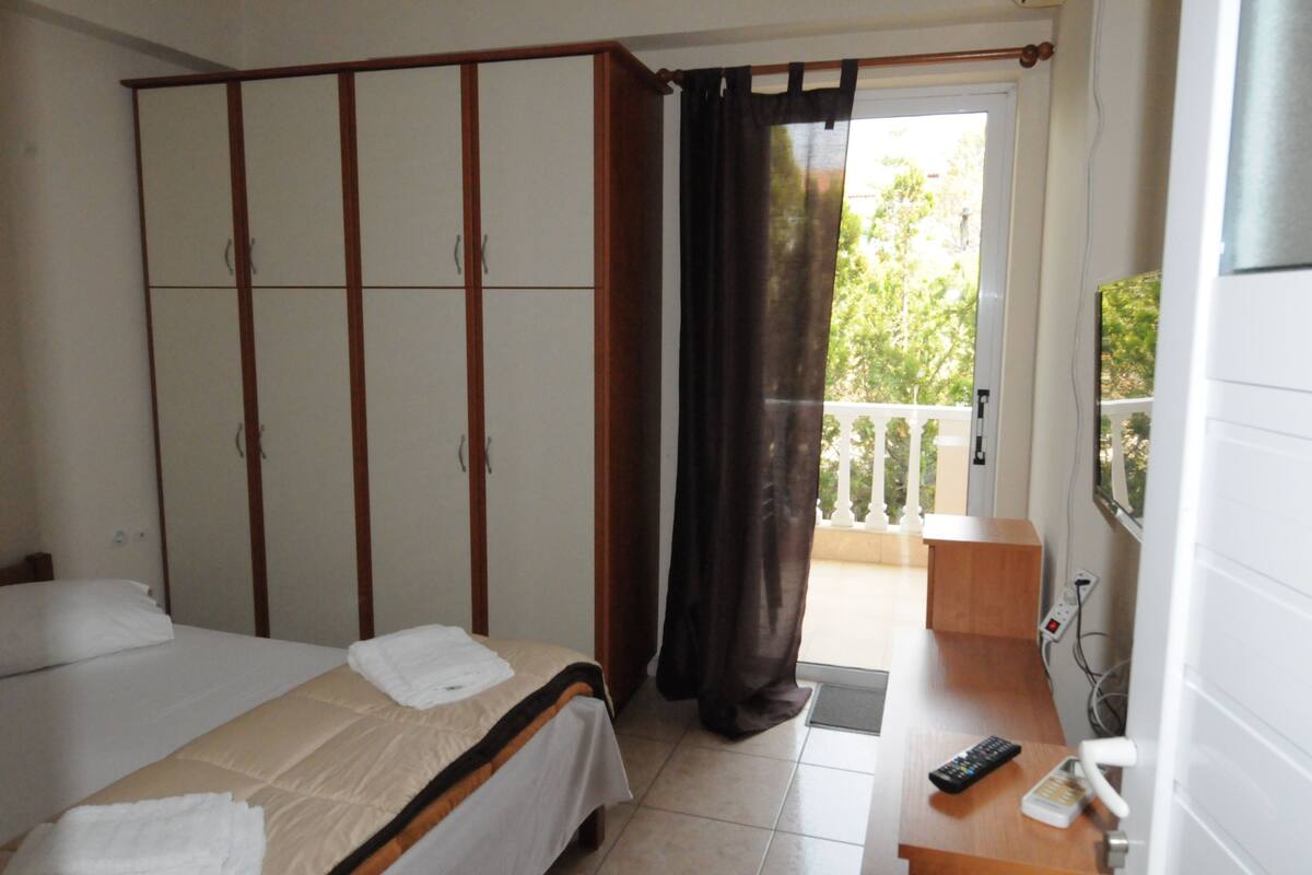 Quarto de hotel com cama, guarda-roupa, varanda e vista, e secretária com comando remoto.