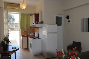 A compact kitchen with a stove, sink, and refrigerator, a dining table with a plant, and a view through a sliding door to an outdoor area.