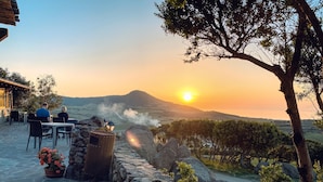 Une terrasse aux murs de pierre, une table avec des chaises et une vue sur le coucher de soleil qui se reflète sur un paysage vallonné.
