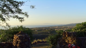Un sentier de pierres mène à un belvédère offrant une vue panoramique, agrémenté de plantes en pot, et d'où l'on aperçoit une ville lointaine et des montagnes.