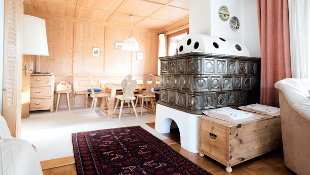 A cozy room with a wooden ceiling, a large stove, a dining area with chairs and a table, and a bed with a white headboard. - Haus Fink (Innsbruck)