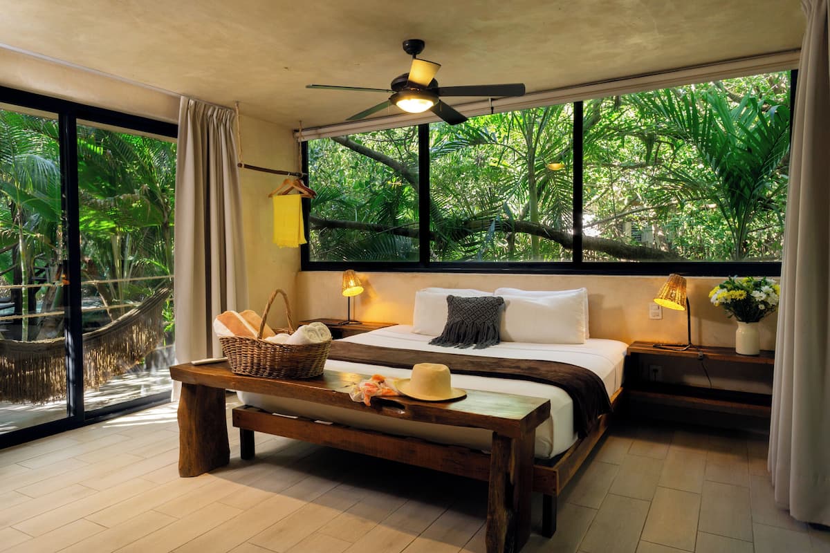 A bedroom with a large window, a bed, a bedside table with a lamp, and a view of greenery outside.