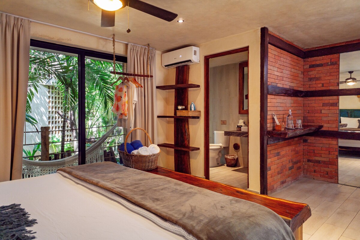 A bedroom with a large bed, a window with a view of greenery, a small bathroom, and a brick accent wall.