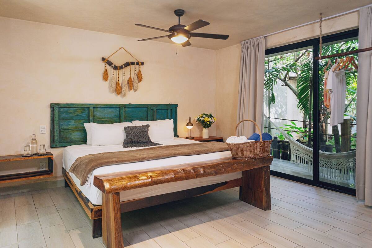 A bedroom with a wooden bed, a ceiling fan, a nightstand, a window with a view of a hammock outside, and a wall-mounted decorative item.
