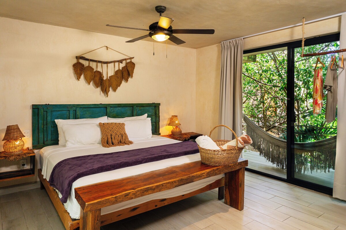 A bedroom with a wooden bed, a ceiling fan, a nightstand with a lamp, and a view of greenery through a large window.