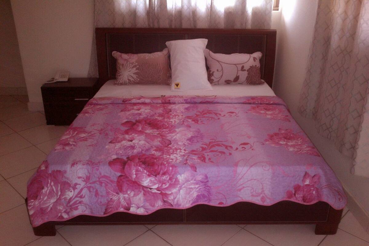 Una cama con colcha floral, cabecera de madera y mesita de noche con un teléfono.