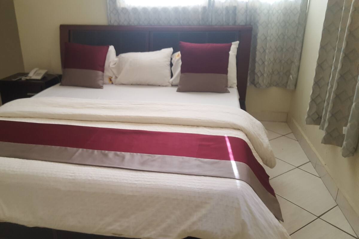 Una cama bien hecha con cabecera, una mesita de noche con teléfono y una ventana con cortinas.