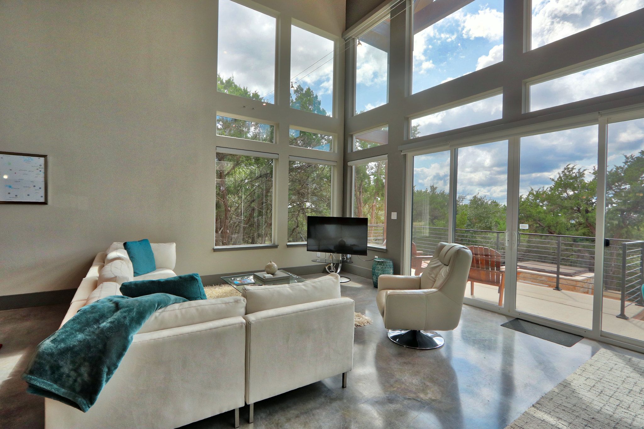 A modern living room with a white sofa, a glass coffee table, and a television. Large windows offer a view of the outdoors.