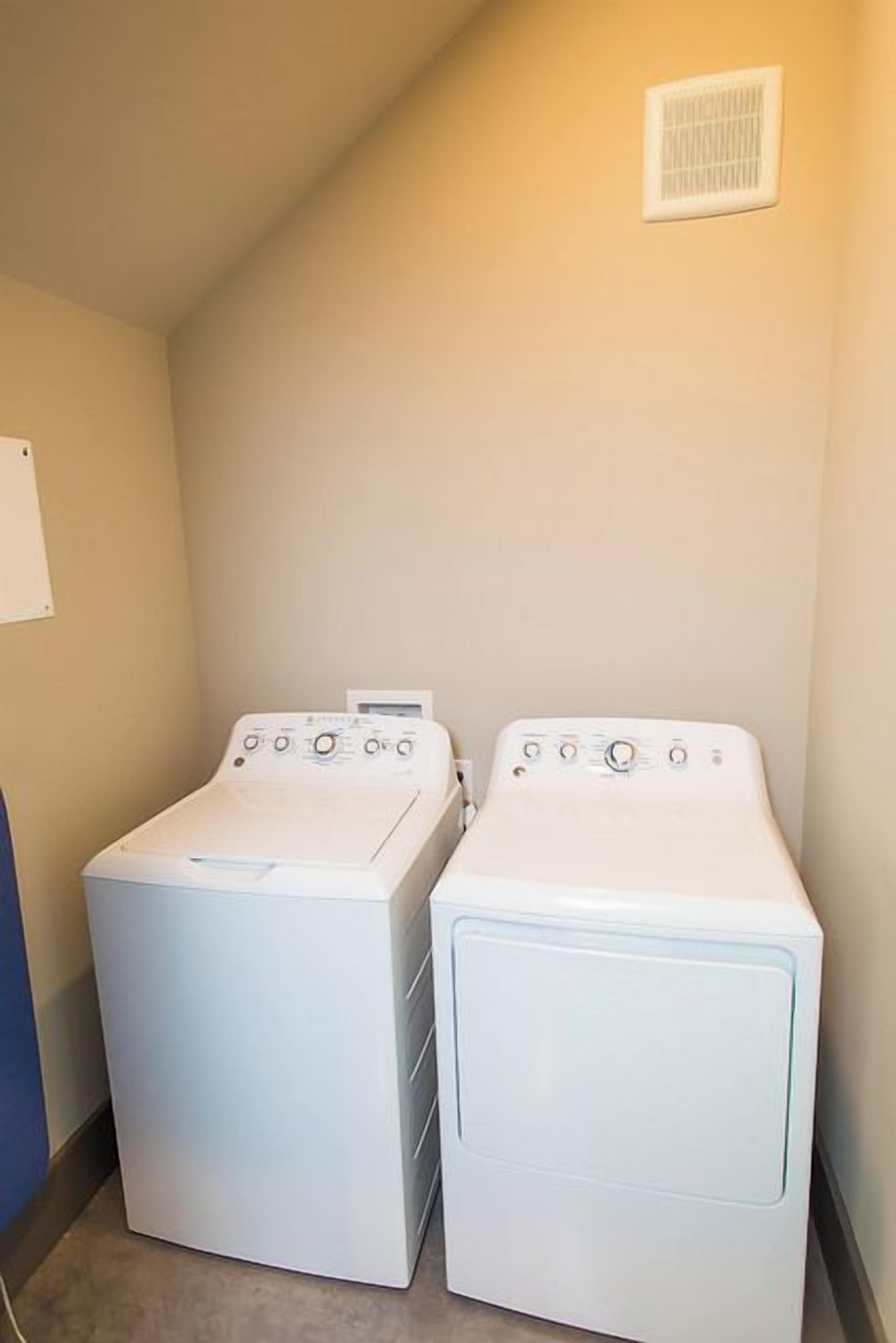 A laundry room with two front-loading washing machines and a vent on the wall.