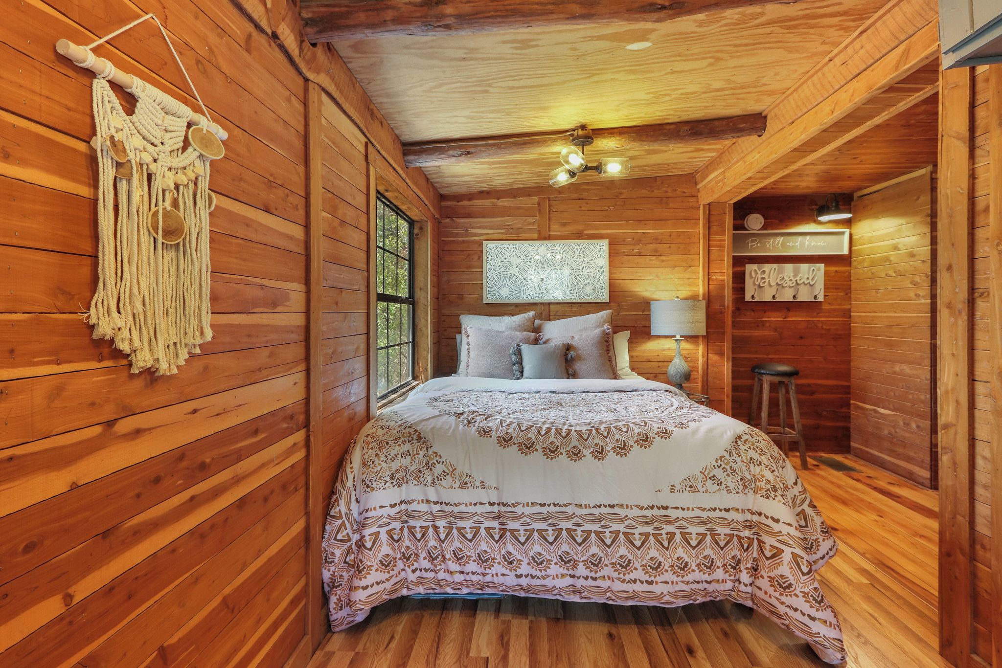 A wooden cabin bedroom with a bed, a macrame wall hanging, a window, and a wall-mounted light fixture.