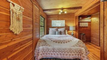 A wooden cabin bedroom with a bed, a macrame wall hanging, a window, and a wall-mounted light fixture.