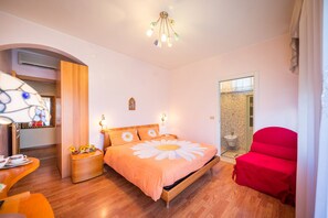 A bedroom with a wooden bed, a nightstand, a red chair, and a bathroom with a shower and toilet. - Hotel Fabrizio (Caorle)
