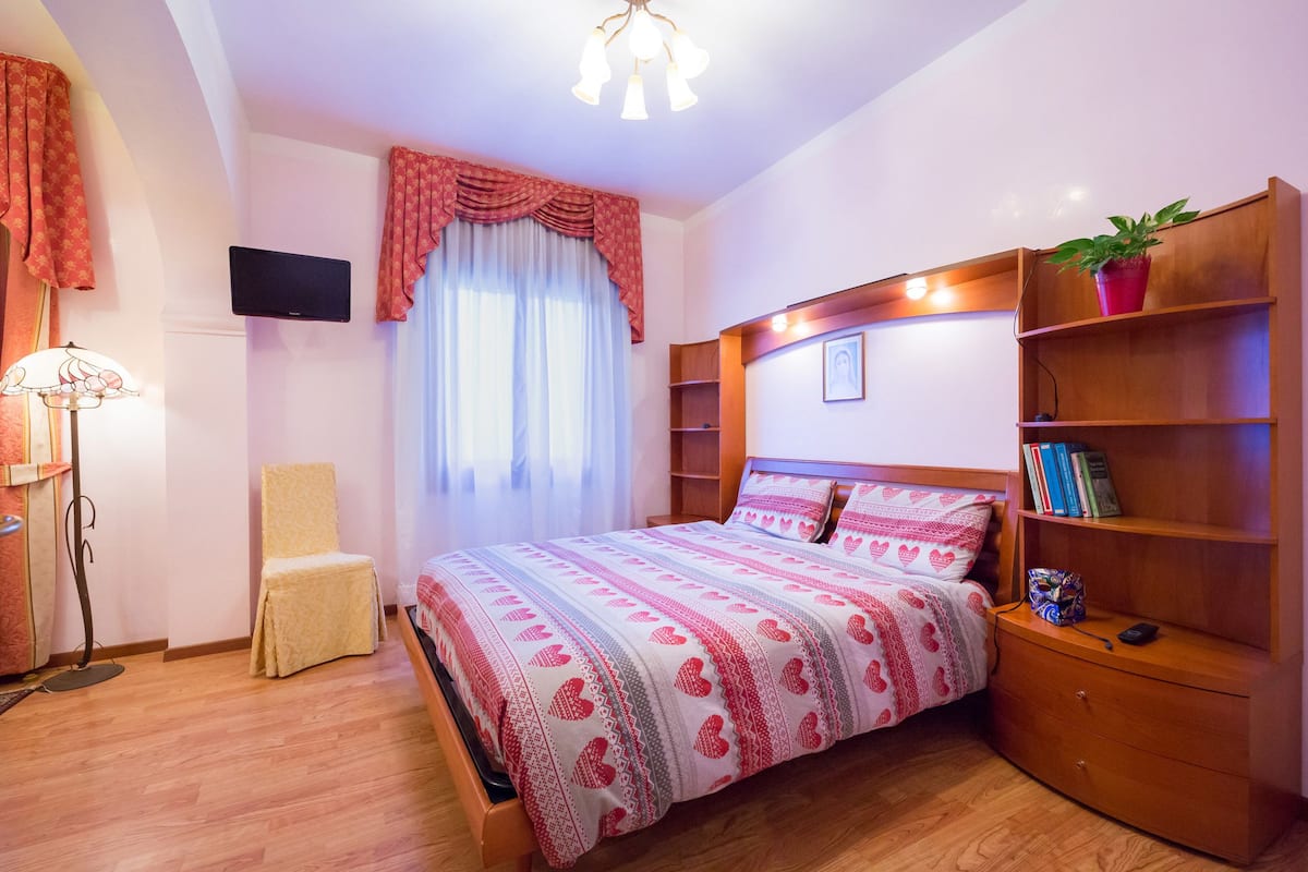 A bedroom with a bed, a chair, a wooden nightstand, a bookshelf, a television, and a window with curtains.