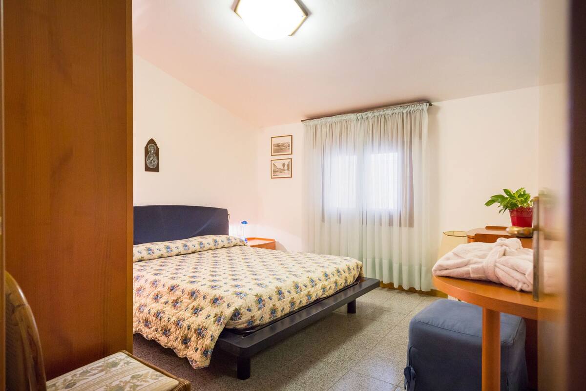 A bedroom with a bed, a bedside table, a chair, a window with curtains, and a potted plant.