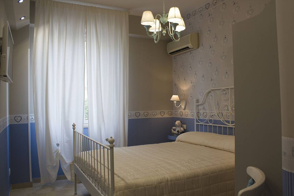 Quarto com cama, lustre, ar condicionado e janela com cortinas.