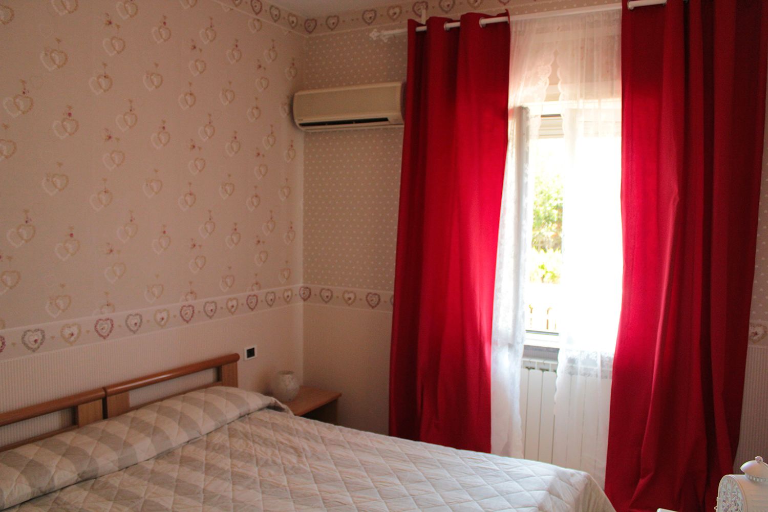 A bedroom with a bed, a nightstand, a window with red curtains, and a wall air conditioner.