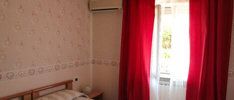 A bedroom with a bed, a nightstand, a window with red curtains, and a wall air conditioner.