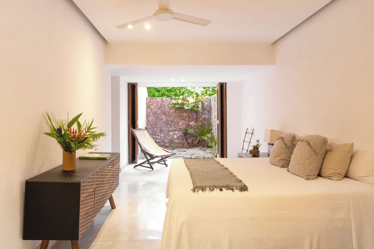 Un dormitorio moderno con cama, mesita de noche, una silla y vistas a un espacio exterior con plantas y una pared de piedra.