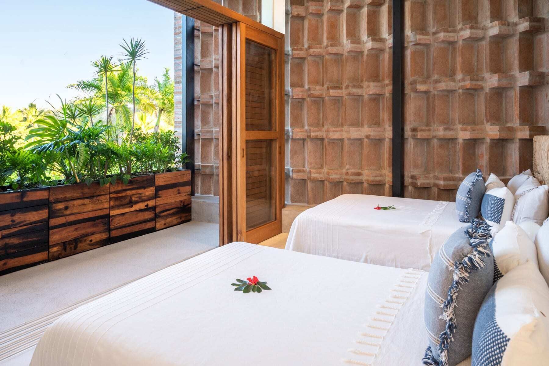 Quarto com duas camas, um banco de madeira com plantas e vista para palmeiras.