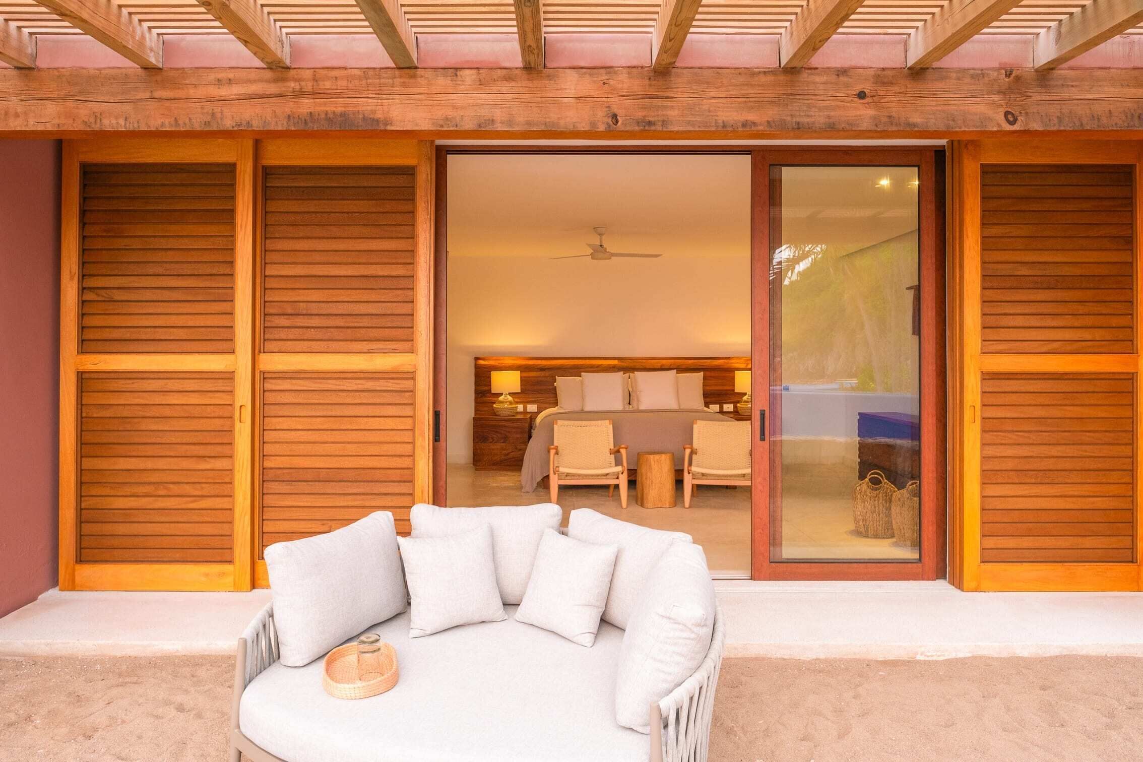 A modern outdoor seating area with a sofa and cushions, a ceiling fan, and a sliding glass door leading to a bedroom with a bed and chairs.