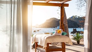 A wooden massage table with a towel and flower, set on a sandy beach with a view of the sea and hills.