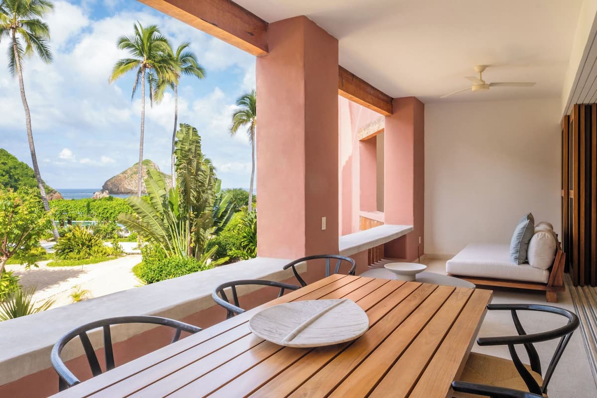 Una mesa de madera con sillas en un balcón con vistas a un paisaje tropical con palmeras y vista al océano.