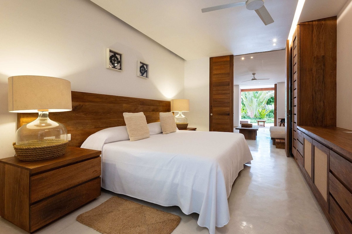 Un dormitorio con cabecera de madera, dos mesitas de noche con lámparas, una cama grande con ropa de cama blanca y vistas a un jardín a través de una puerta abierta.