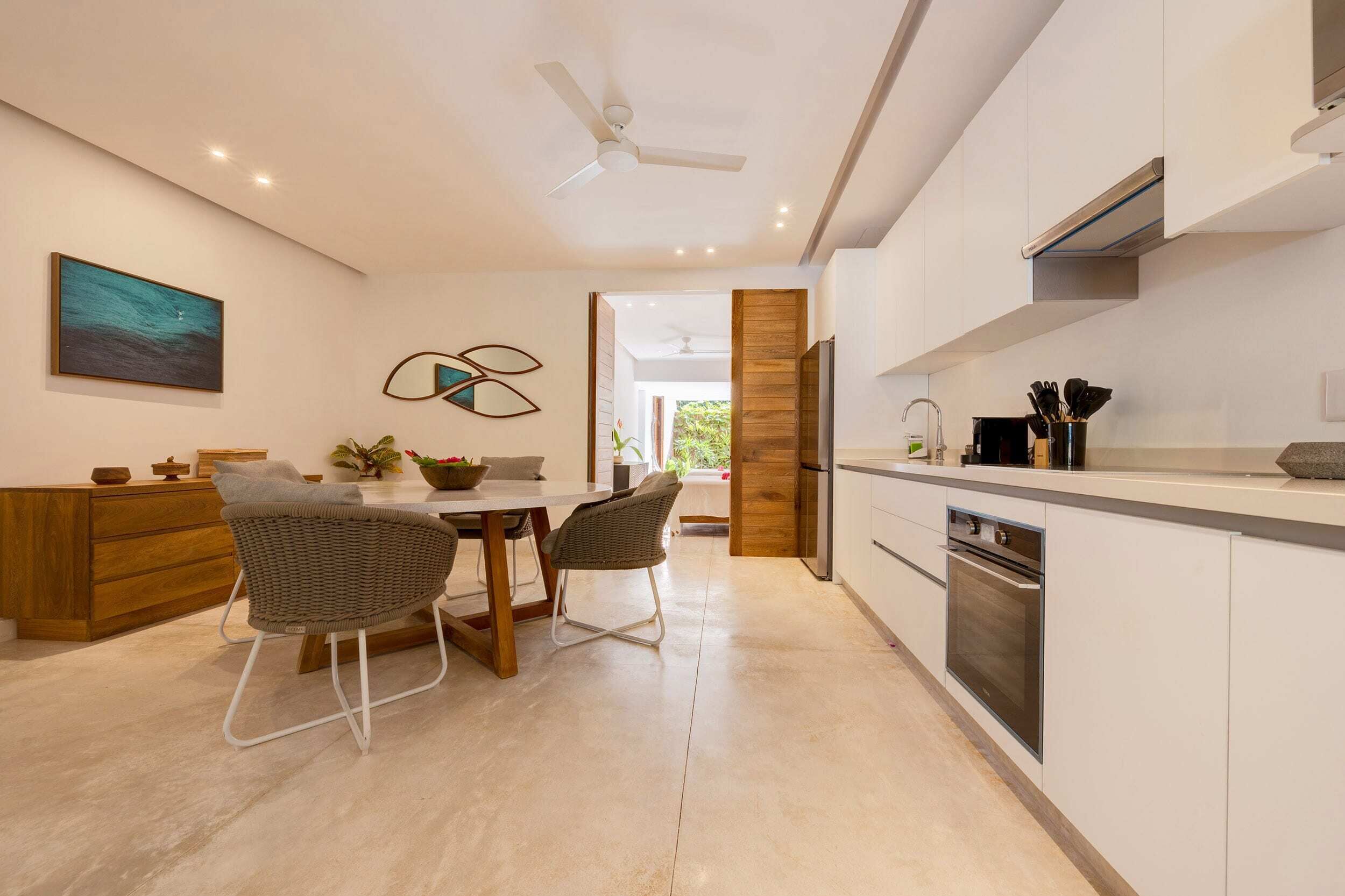 Una cocina moderna con armarios blancos, horno empotrado y comedor con sillas de mimbre.