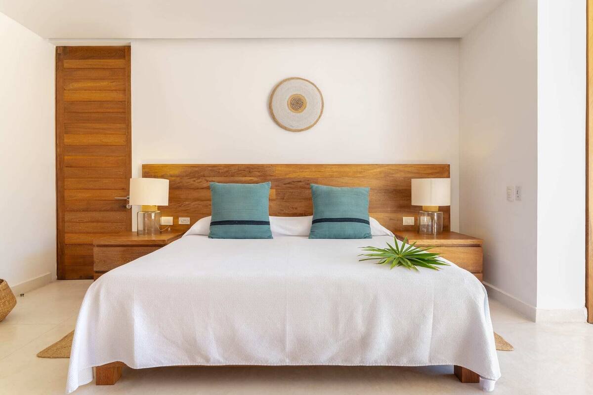 Una cama bien hecha con una colcha blanca y dos almohadas color turquesa, flanqueada por dos mesitas de noche con lámparas, en una habitación con cabecera de madera y un pequeño adorno en la pared.
