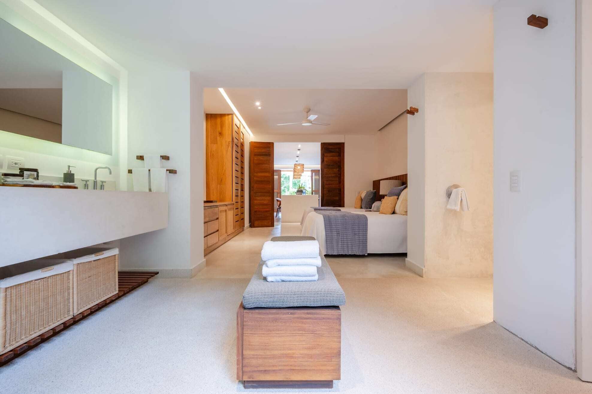 Una habitación de hotel moderna con un espejo grande, armarios de madera, una cama con almohadas, una mesita de noche con toallas y vistas al exterior.